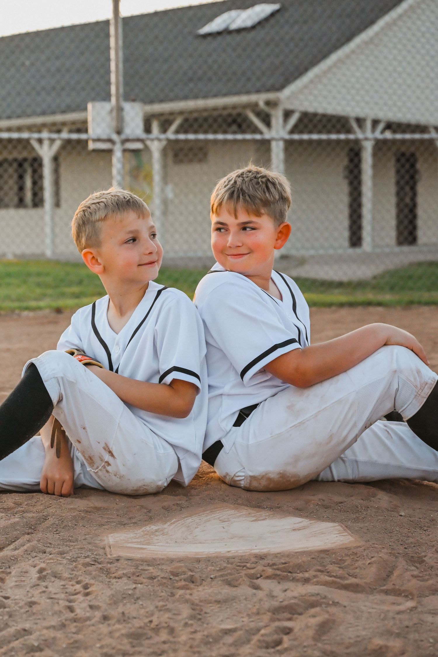 Two young boys in white baseball uniforms sitting back-to-back on a baseball diamond.