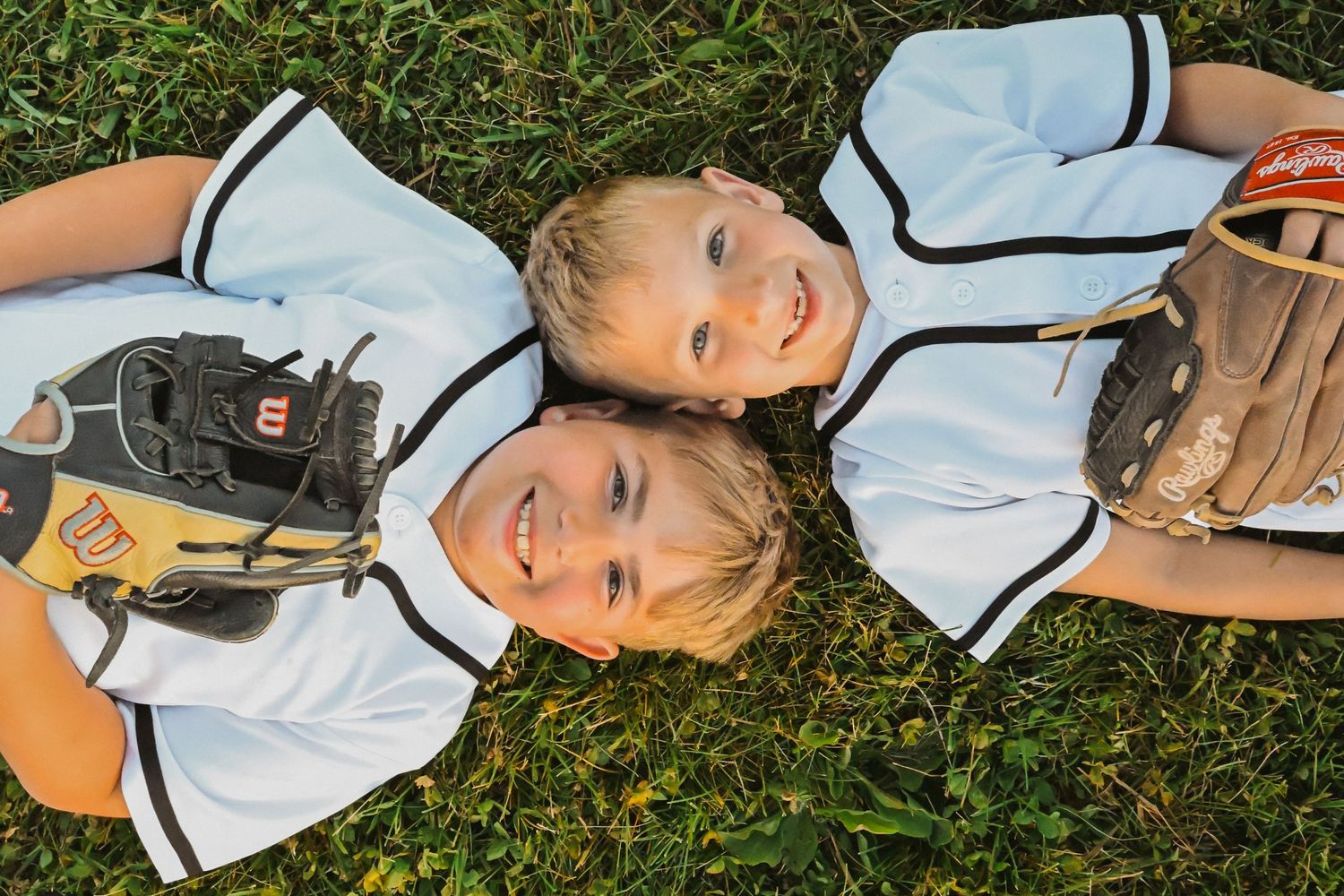 Two young baseball players lying in grass smiling, wearing white uniforms with gloves.
