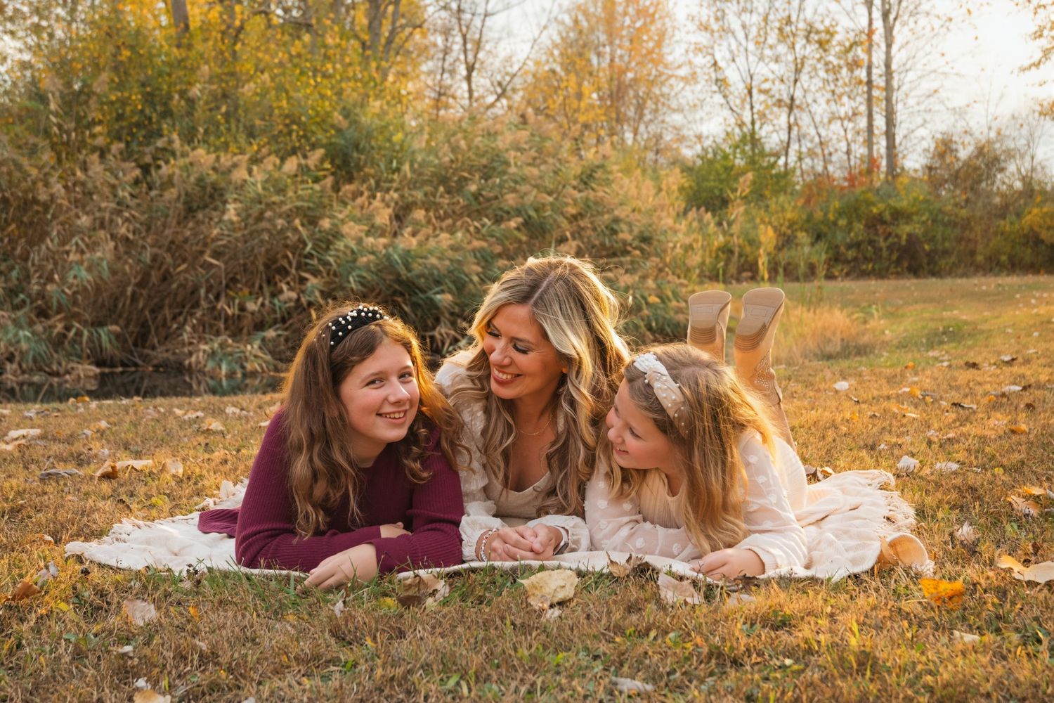 Blonde mother lying on blanket in autumn park, laughing with two daughters wearing floral crowns in fall foliage.