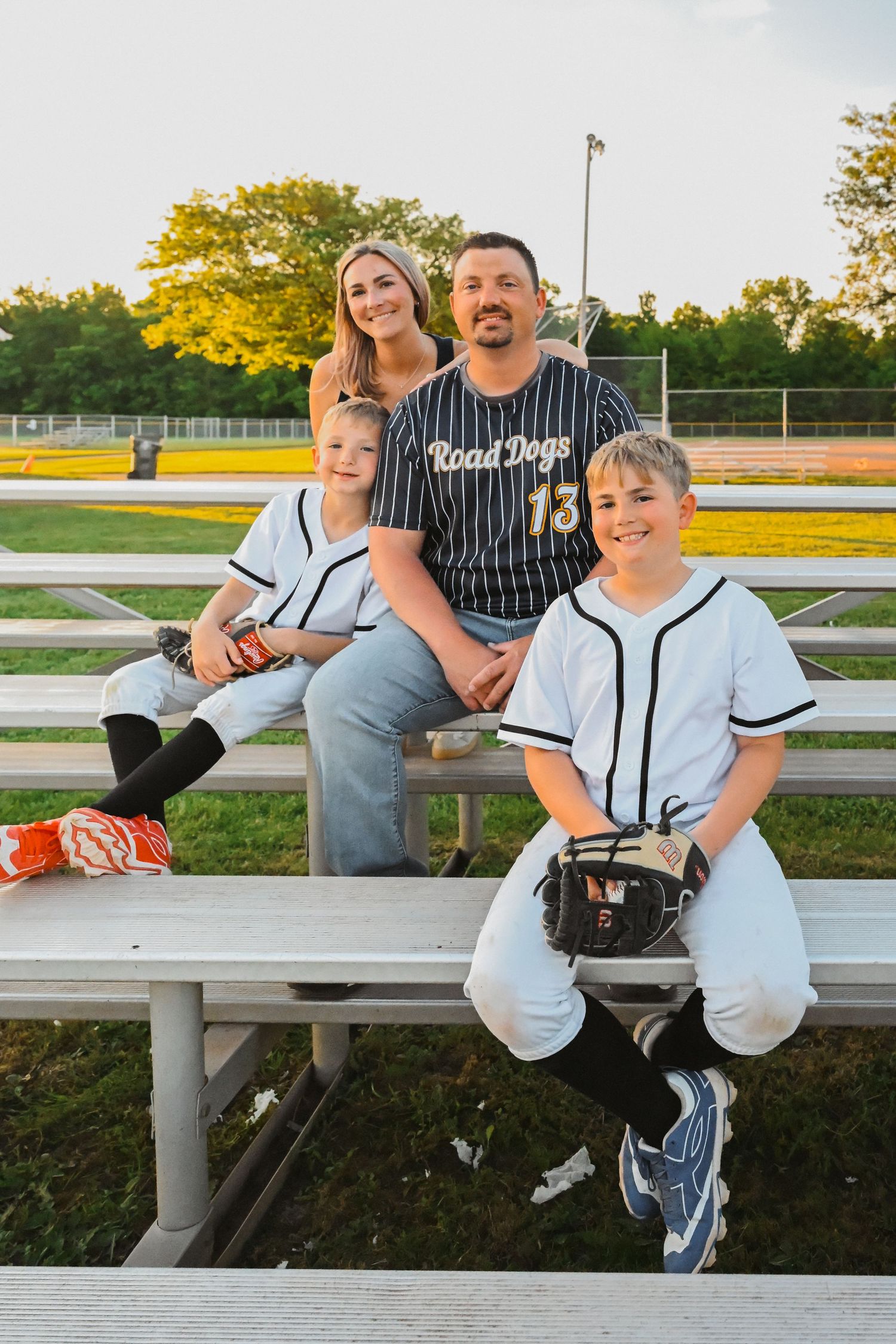 Family of four posing on bleachers at baseball field, two boys in white uniforms.