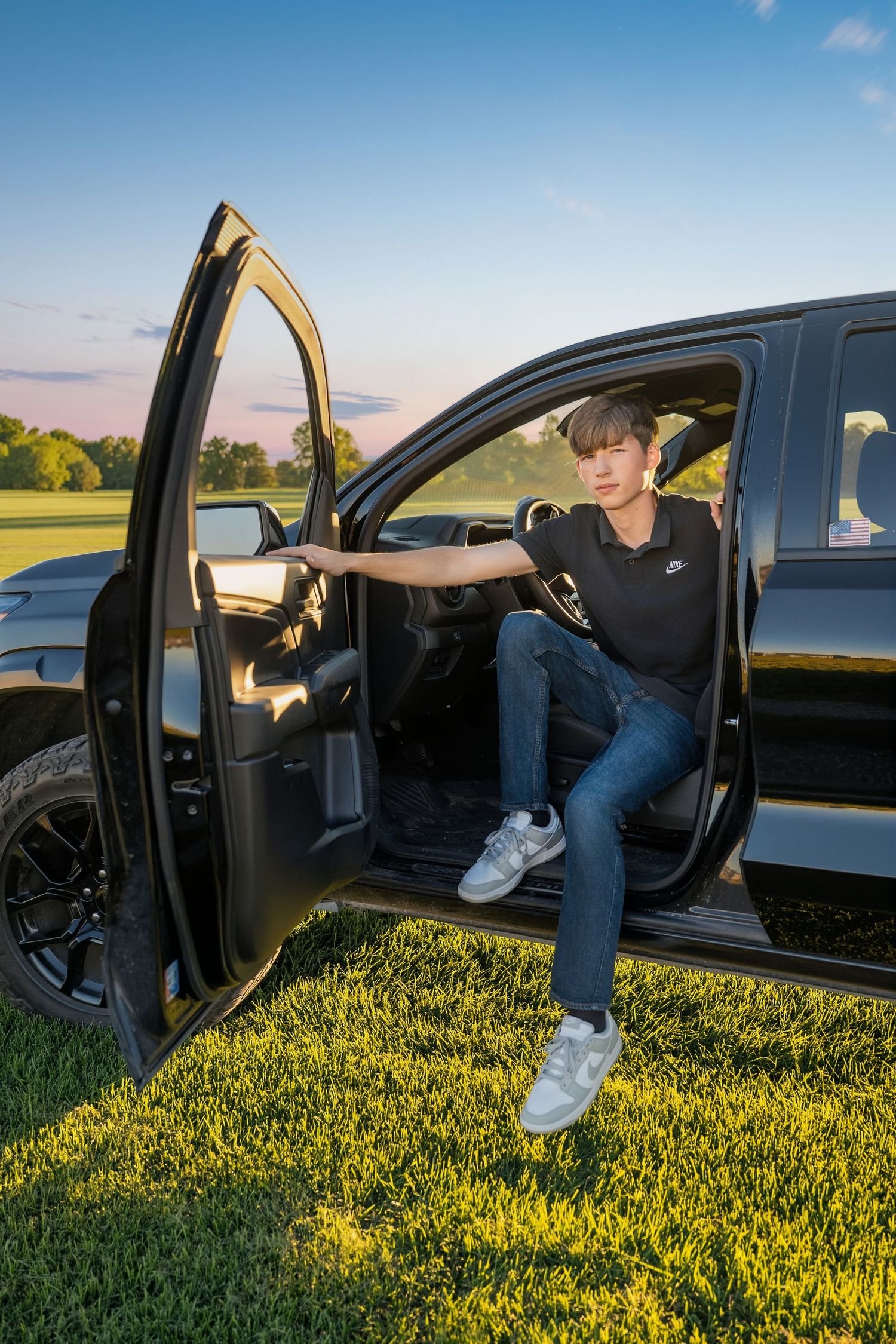 Teen boy in Nike shirt and jeans sits in open door of black truck on green grass at sunset.