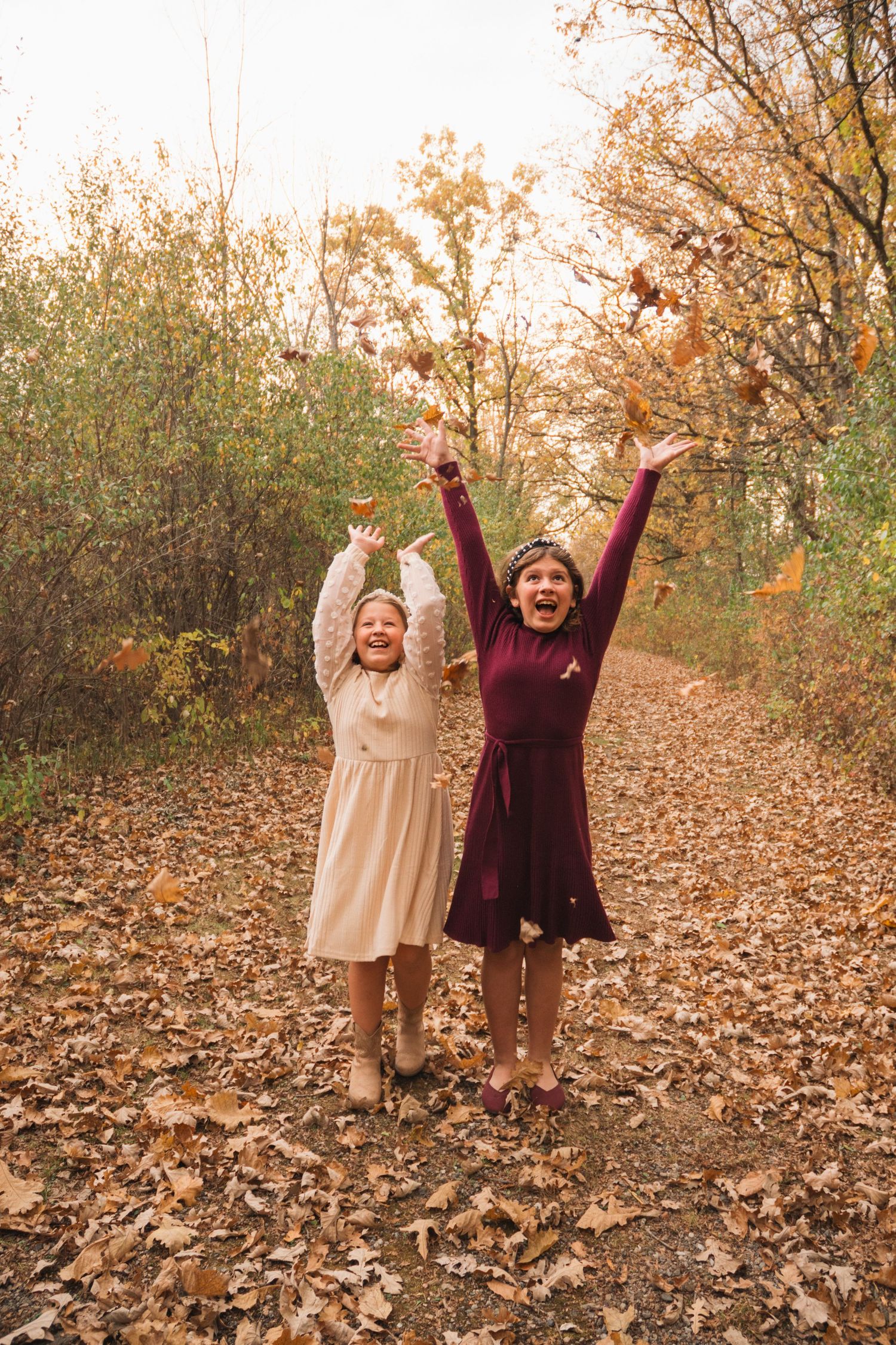 Two girls joyfully tossing autumn leaves on a forest path covered in fallen foliage.
