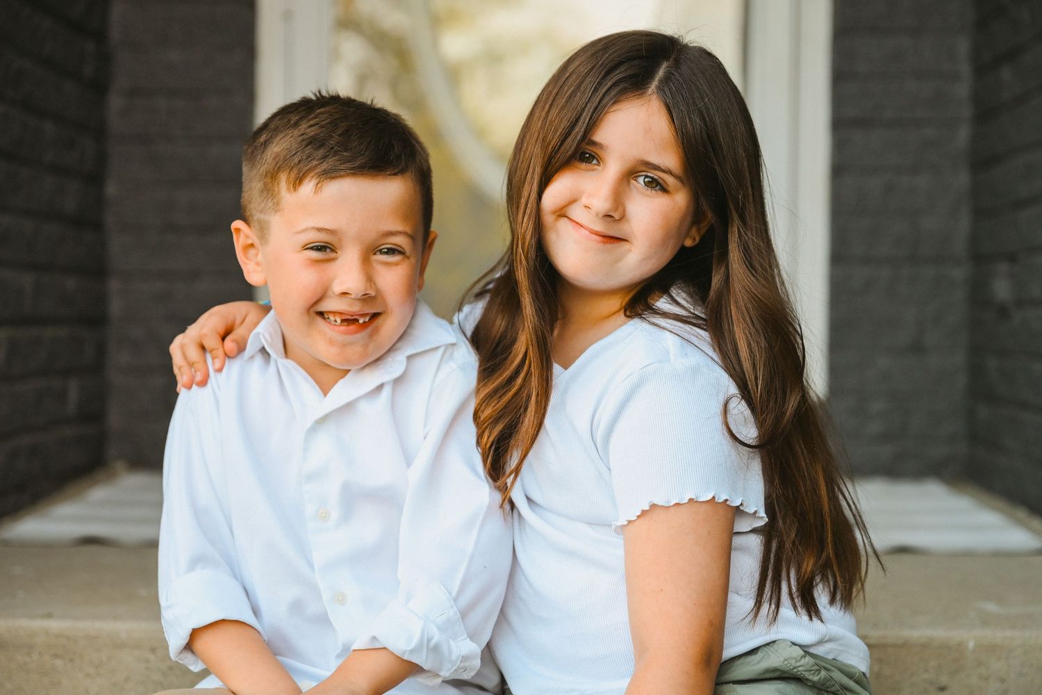 Two happy children in white shirts sitting together on porch steps smiling at camera.