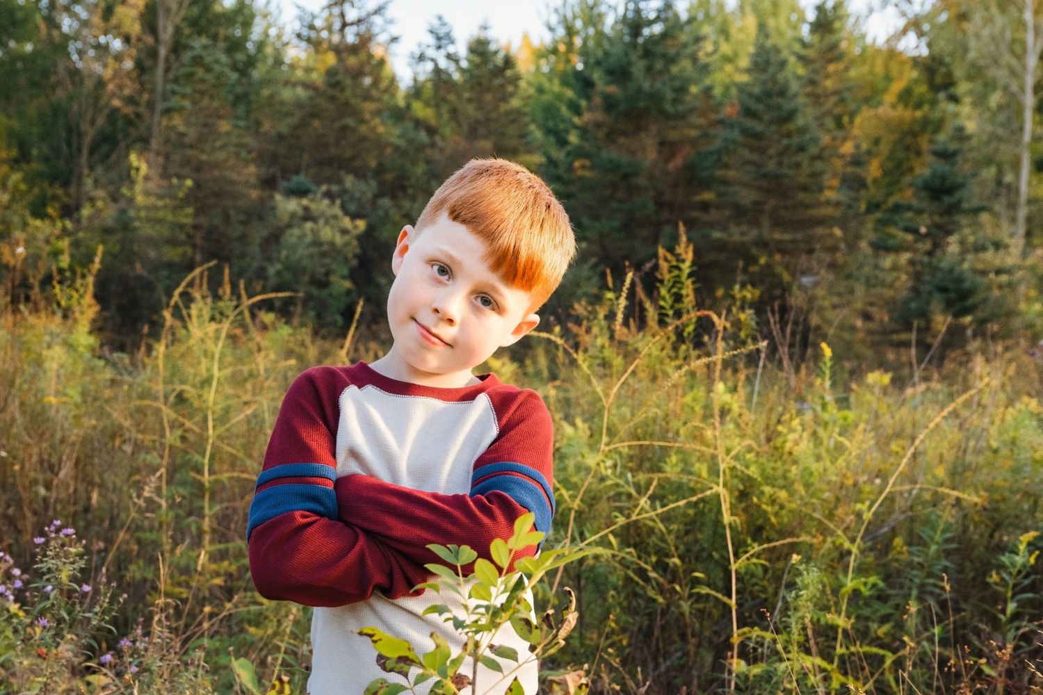 Red-haired boy with arms crossed smiling in a lush green field with trees in the background on a sunny day.