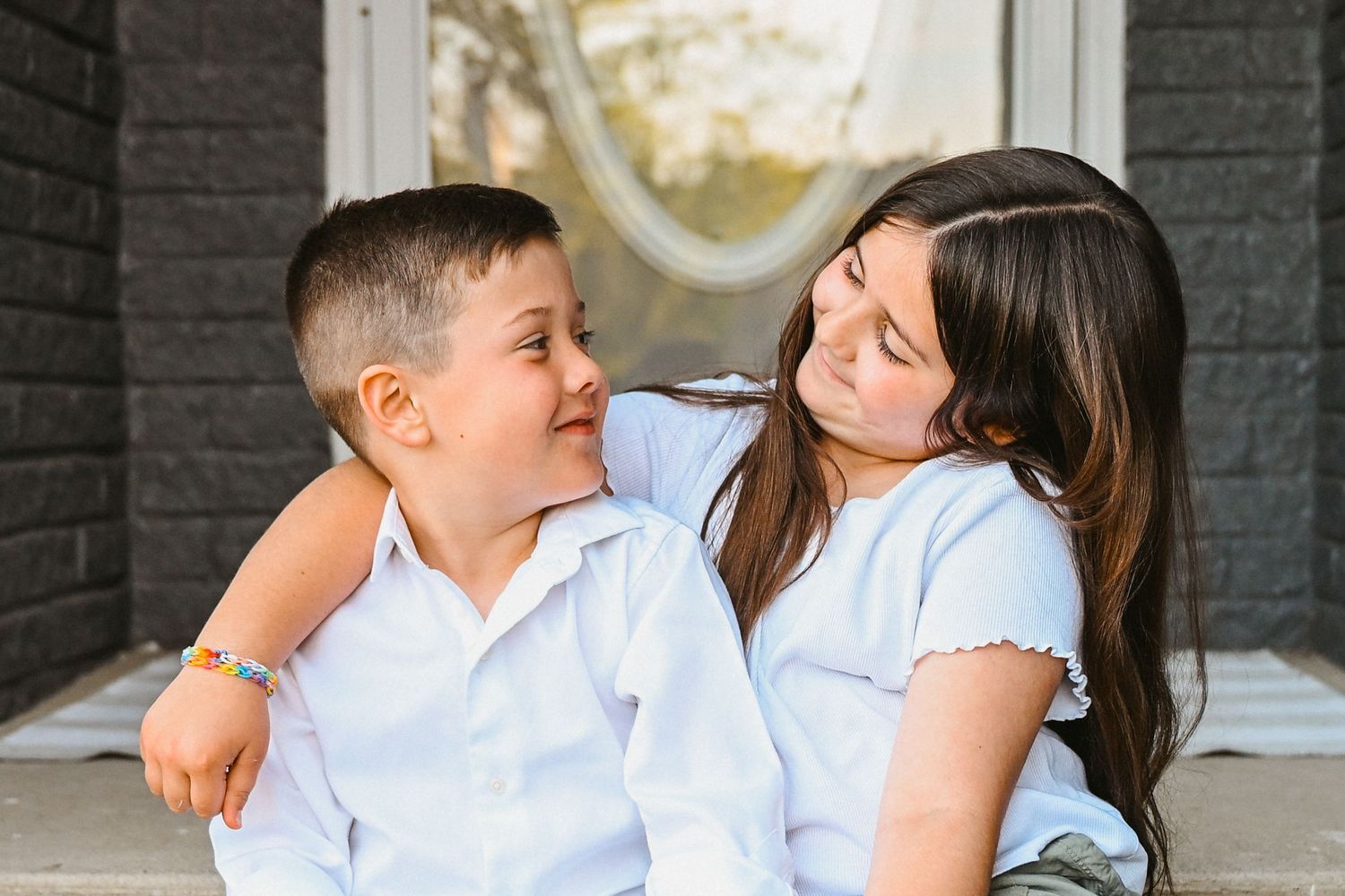 Young girl and boy in white shirts sitting on porch steps smiling at each other.