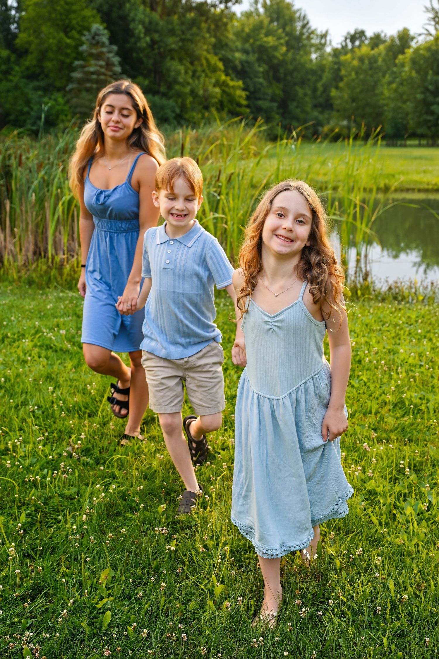Mother walking with two happy children in blue outfits near a pond in nature.