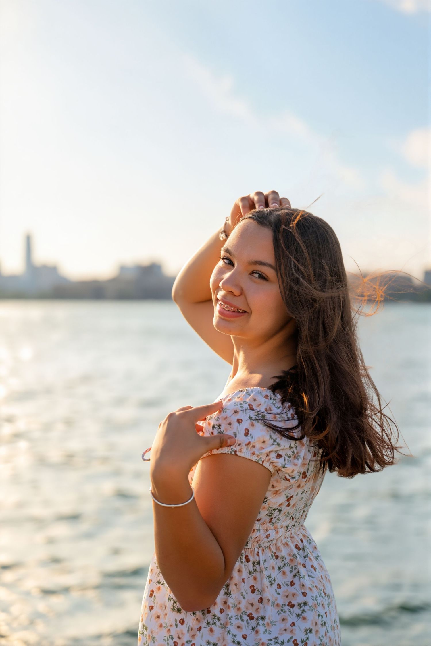 Young woman in floral dress poses by sunlit waterfront, hand in hair, smiling over shoulder.