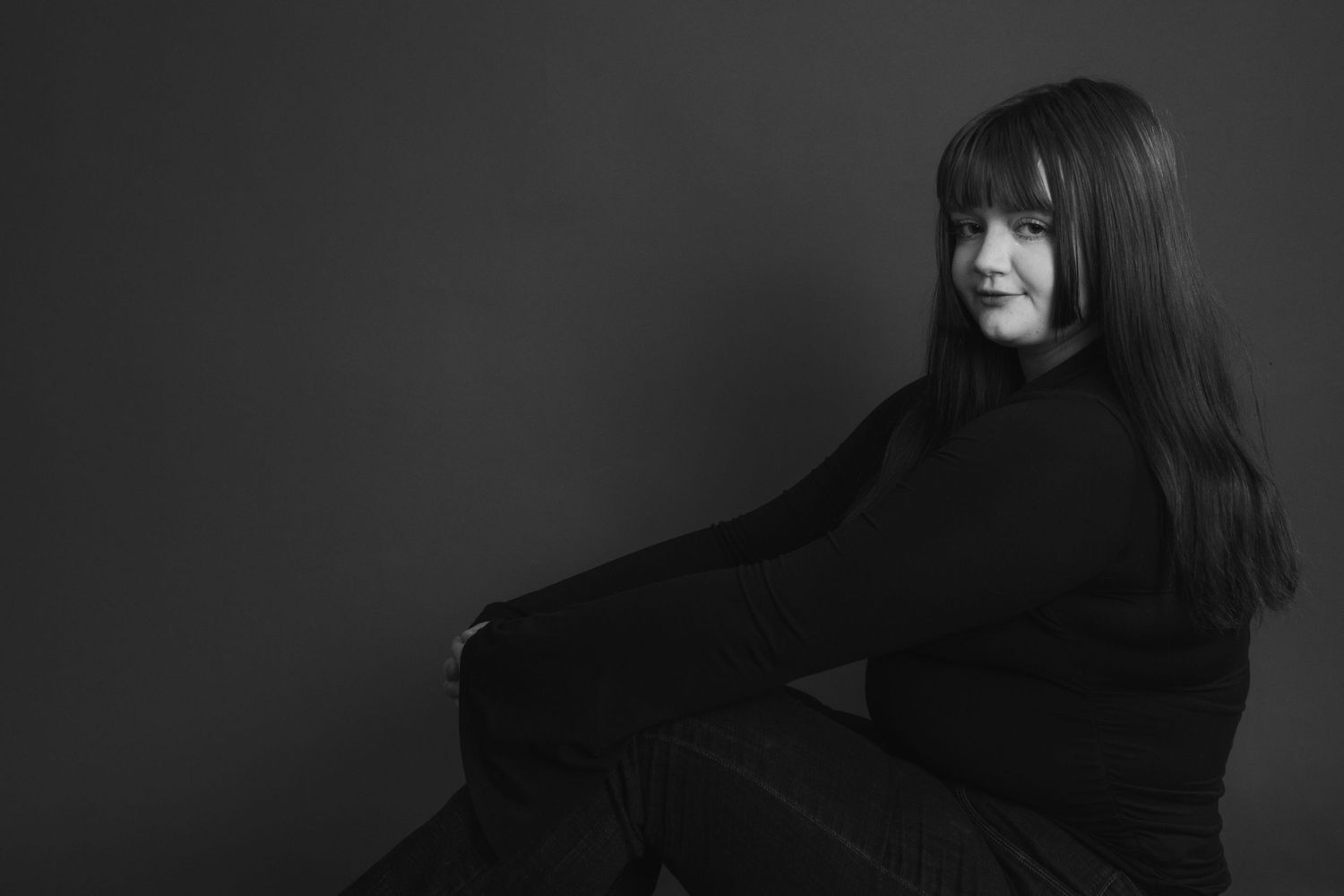 A black and white studio portrait of a young woman with bangs sitting gracefully in a black outfit.
