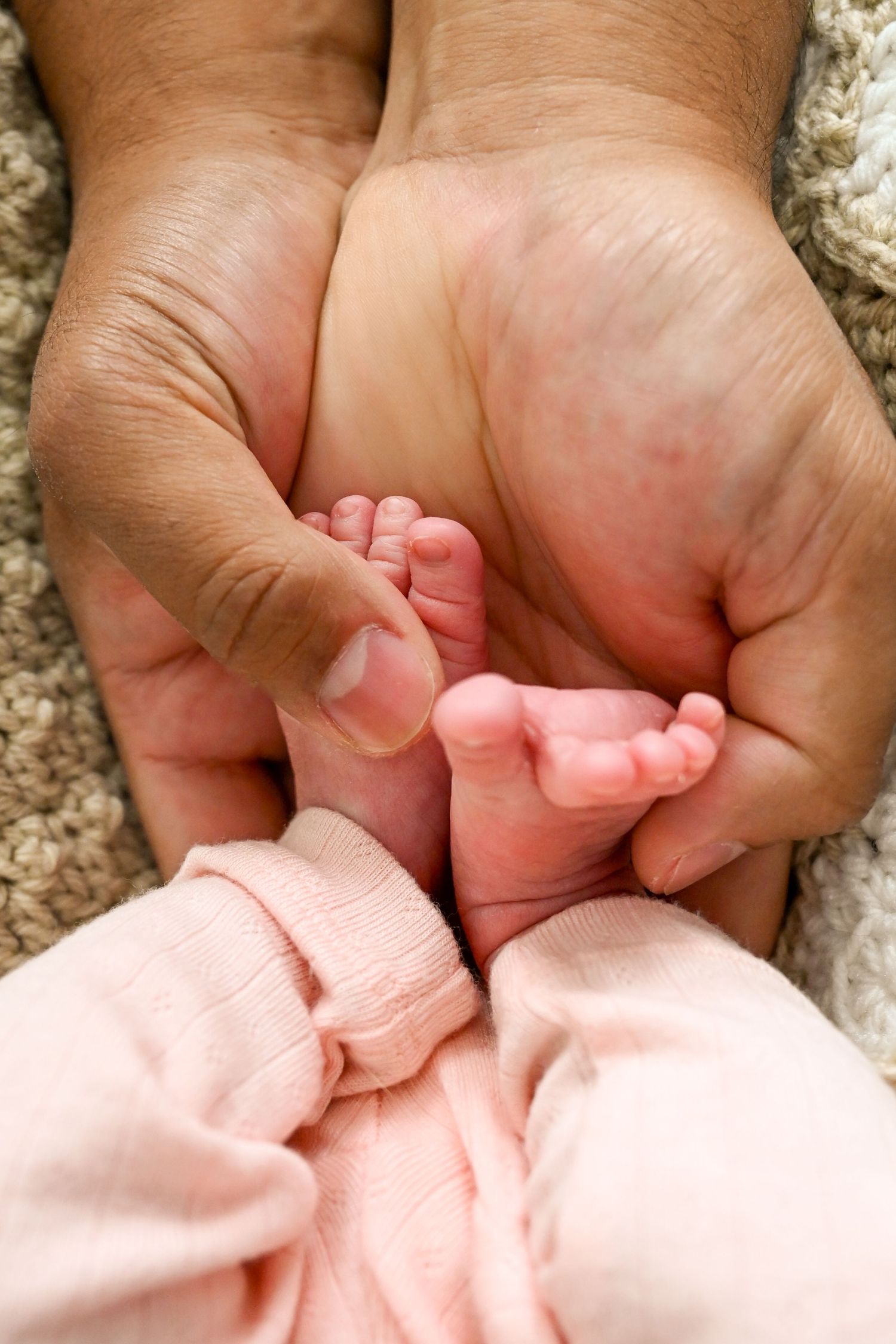 Two adult hands gently cradling the tiny feet of a newborn baby in pink outfit.