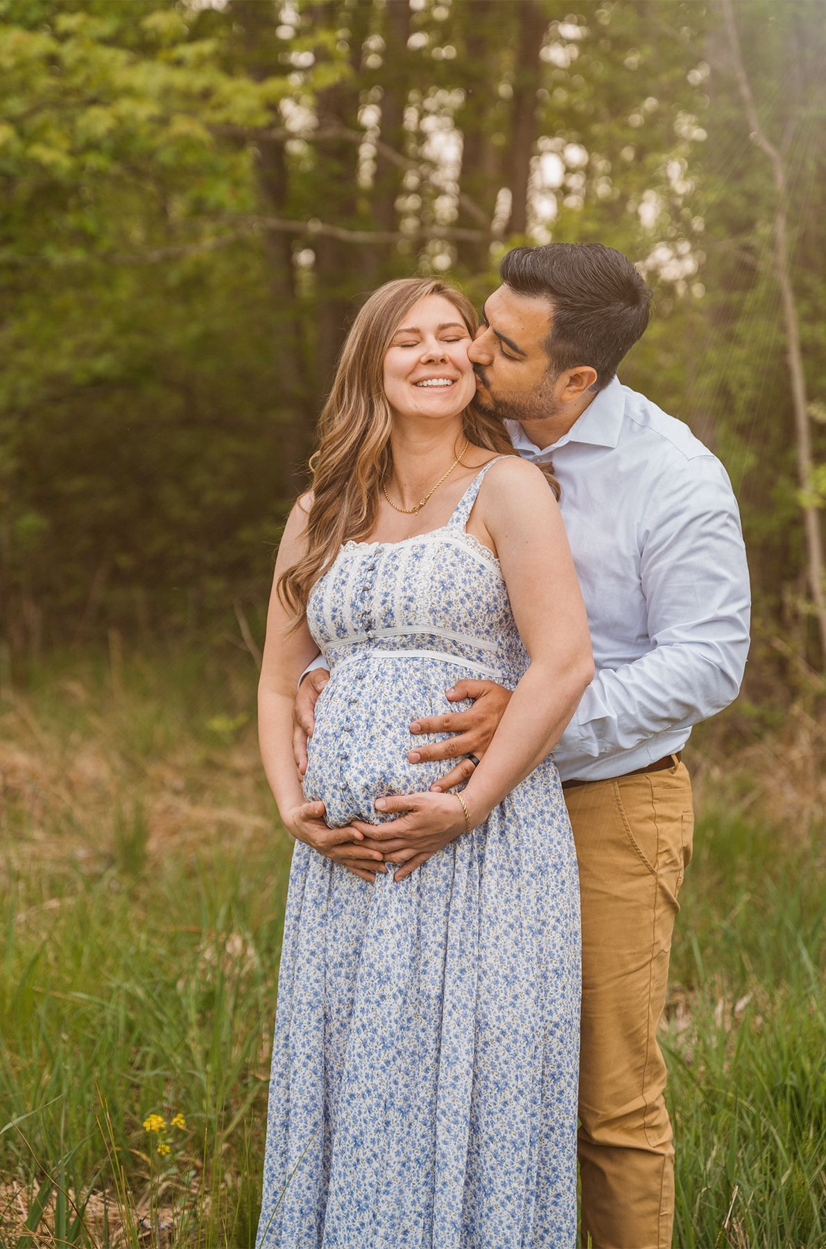 A pregnant woman in a floral dress smiles as her partner kisses her cheek outdoors.