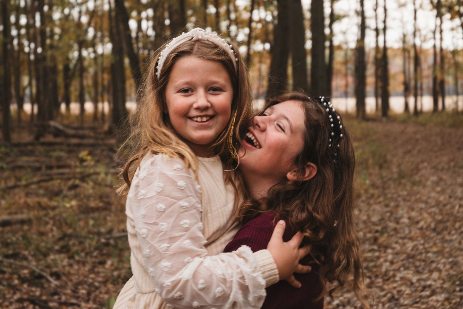 Two young sisters laugh and embrace joyfully together in a sunlit autumn forest setting.