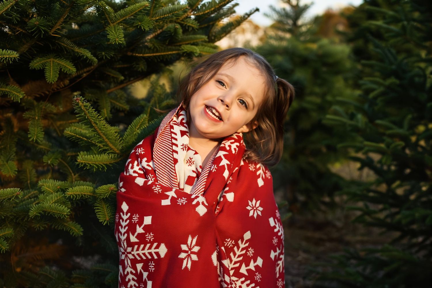 A happy young girl wrapped in a red Nordic pattern blanket smiling at a Christmas tree farm outdoors.