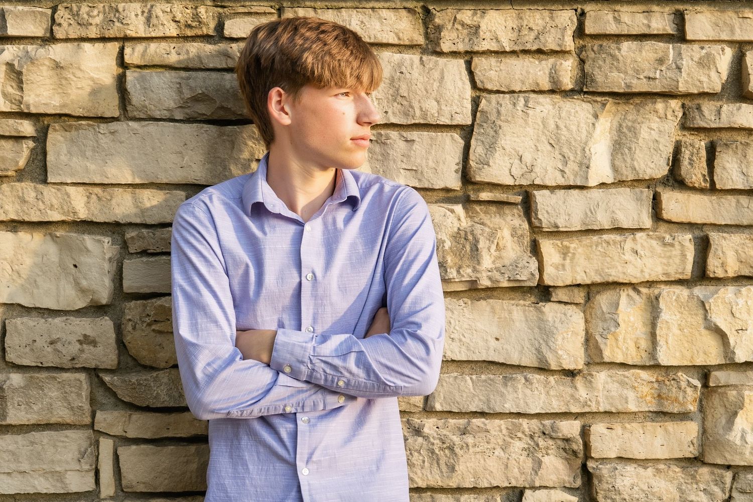 Young man in blue shirt leaning against stone wall with arms crossed, looking sideways in natural light.
