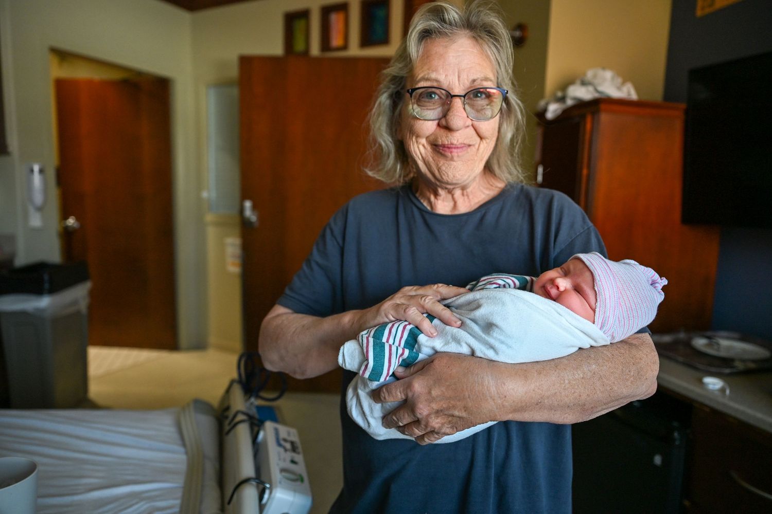 Elderly woman with glasses holding a swaddled newborn baby in a hospital room.