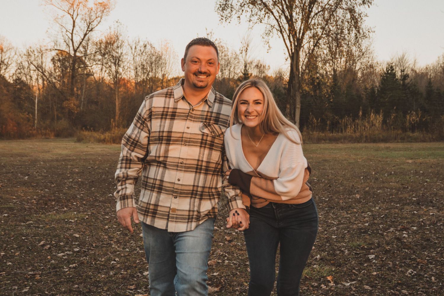 A smiling couple holds hands in a golden autumn field, man in plaid flannel and woman in cream top.