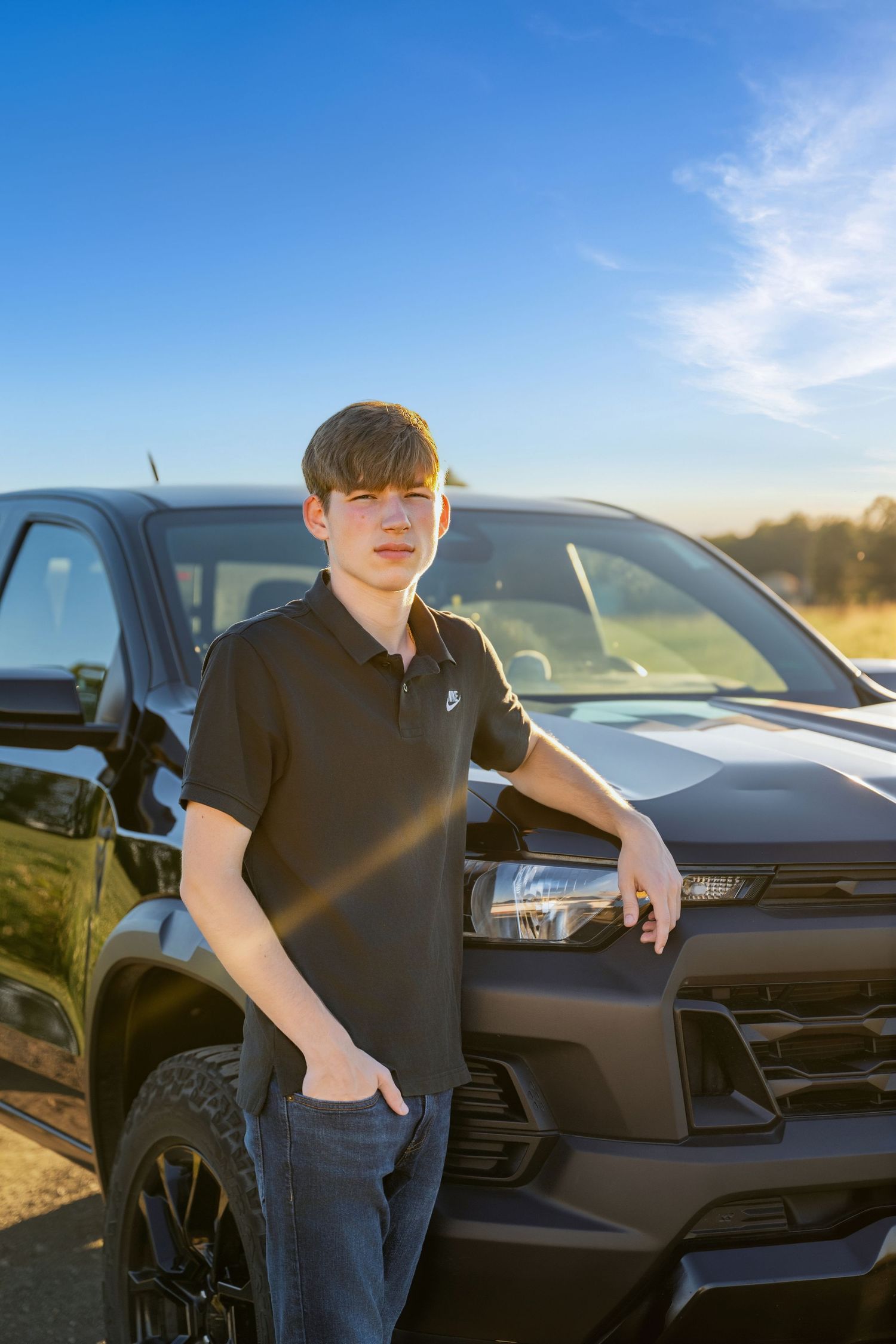 Teenage boy in a black polo shirt leaning on the hood of a black pickup truck outdoors.