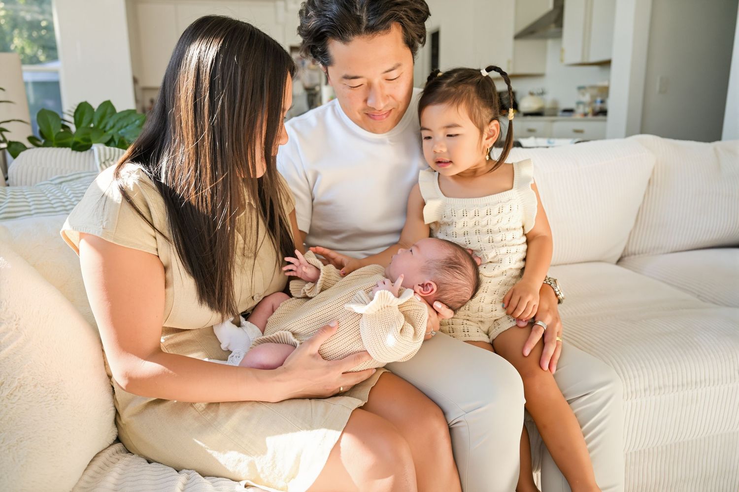 Asian parents and two children sharing tender moment on white couch, admiring their newborn baby together at home.