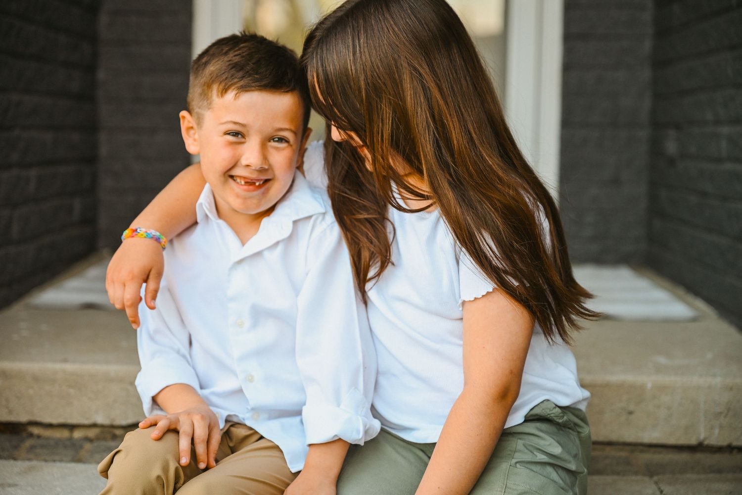 Smiling young boy in white shirt being hugged by older sister on front porch steps.