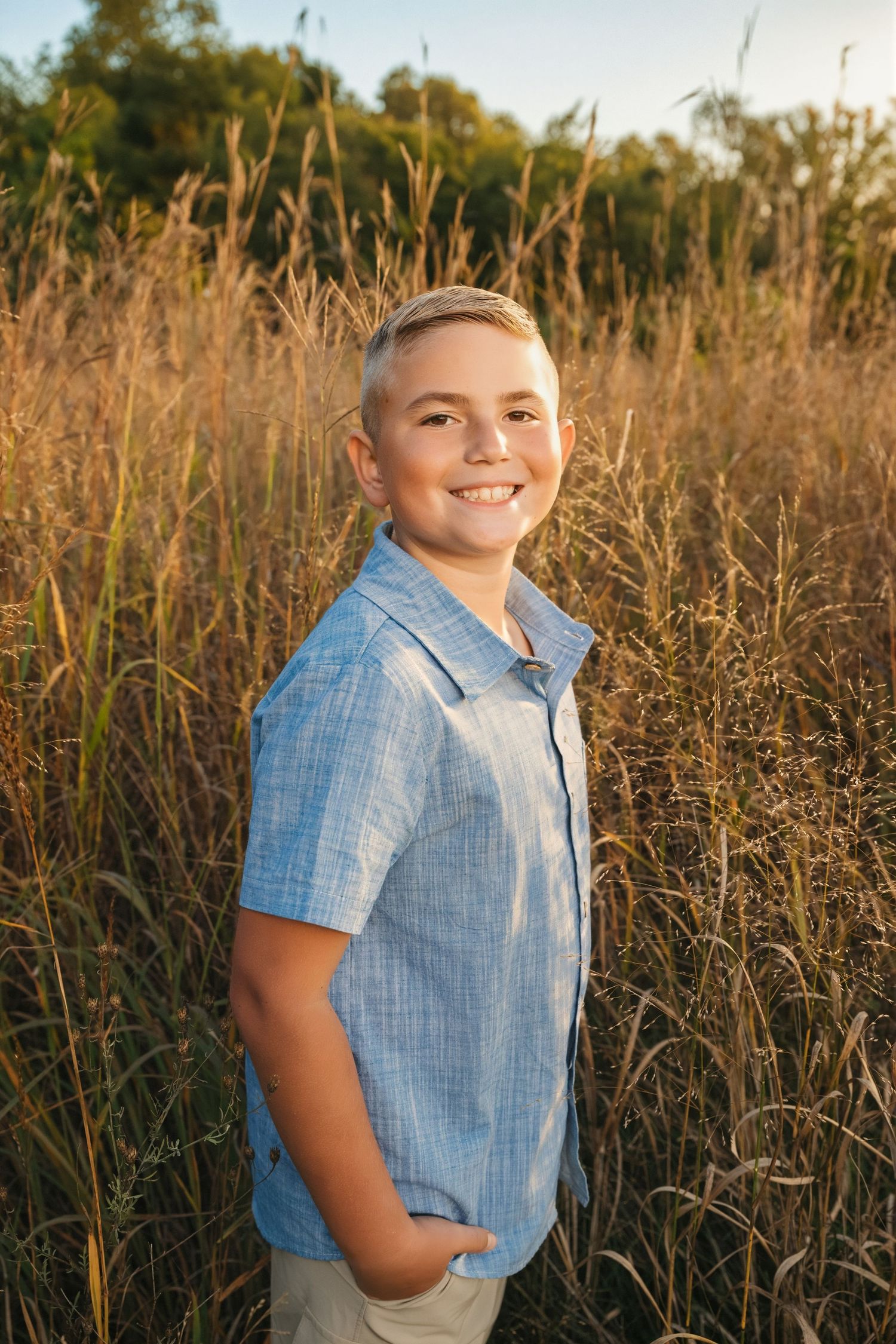 Smiling boy in blue shirt standing in golden tall grass field during sunset.