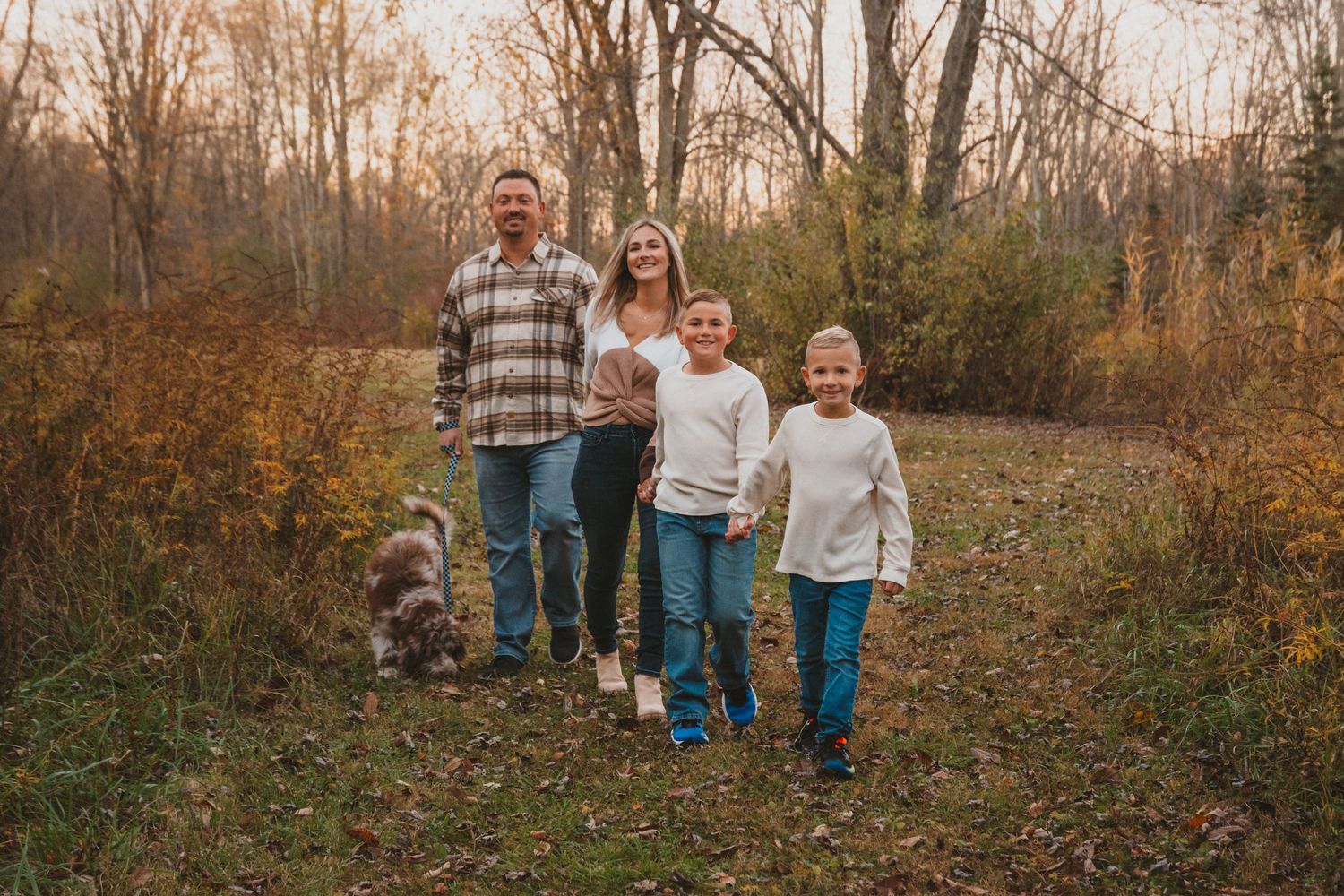 A happy family of four walks together along an autumn forest path with their small dog.