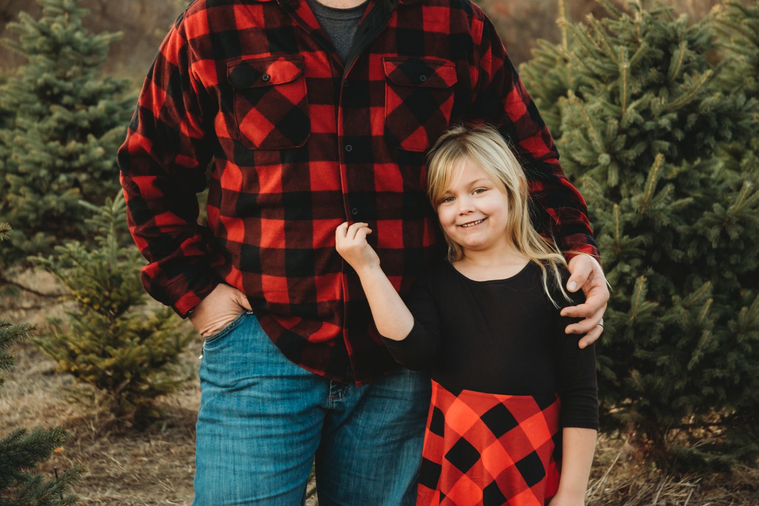 Father and daughter in matching red buffalo plaid outfits smiling at a Christmas tree farm.