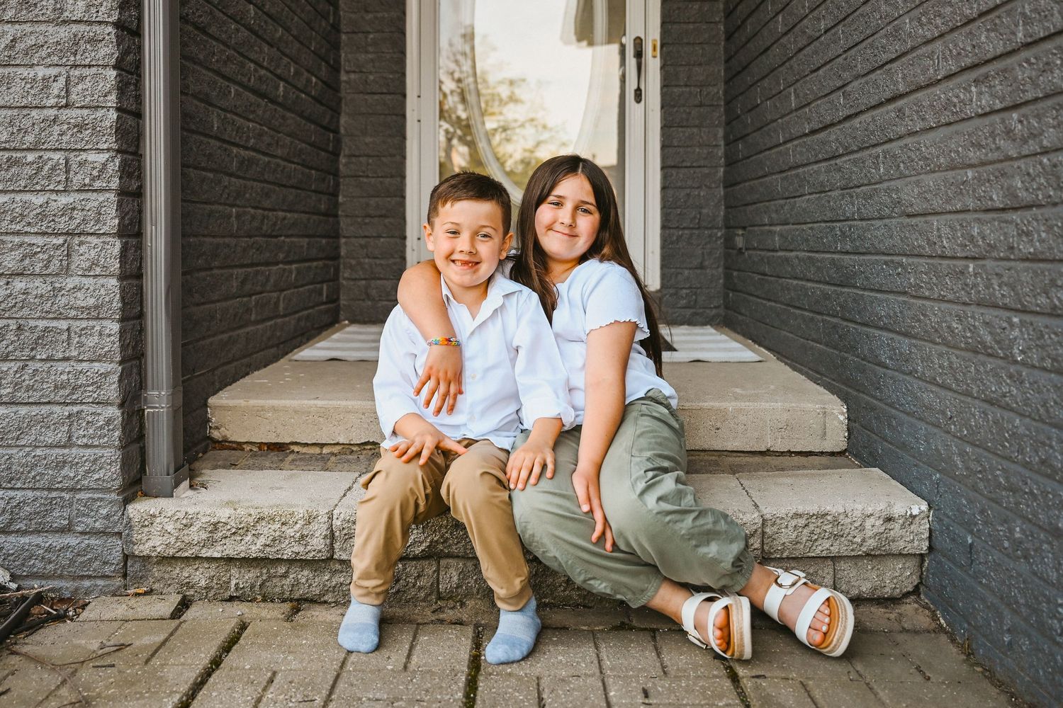 Two smiling children sit on front porch steps holding each other, wearing white shirts in a warm outdoor setting.