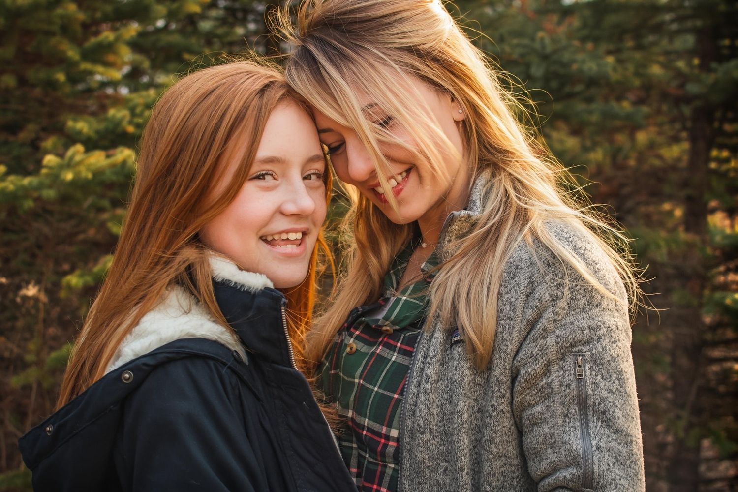 Two smiling women with red and blonde hair lean foreheads together outdoors in autumn forest setting.