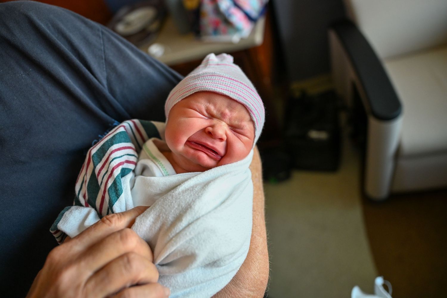 Newborn baby crying wrapped in white swaddle blanket wearing pink striped hat being held in hospital room.