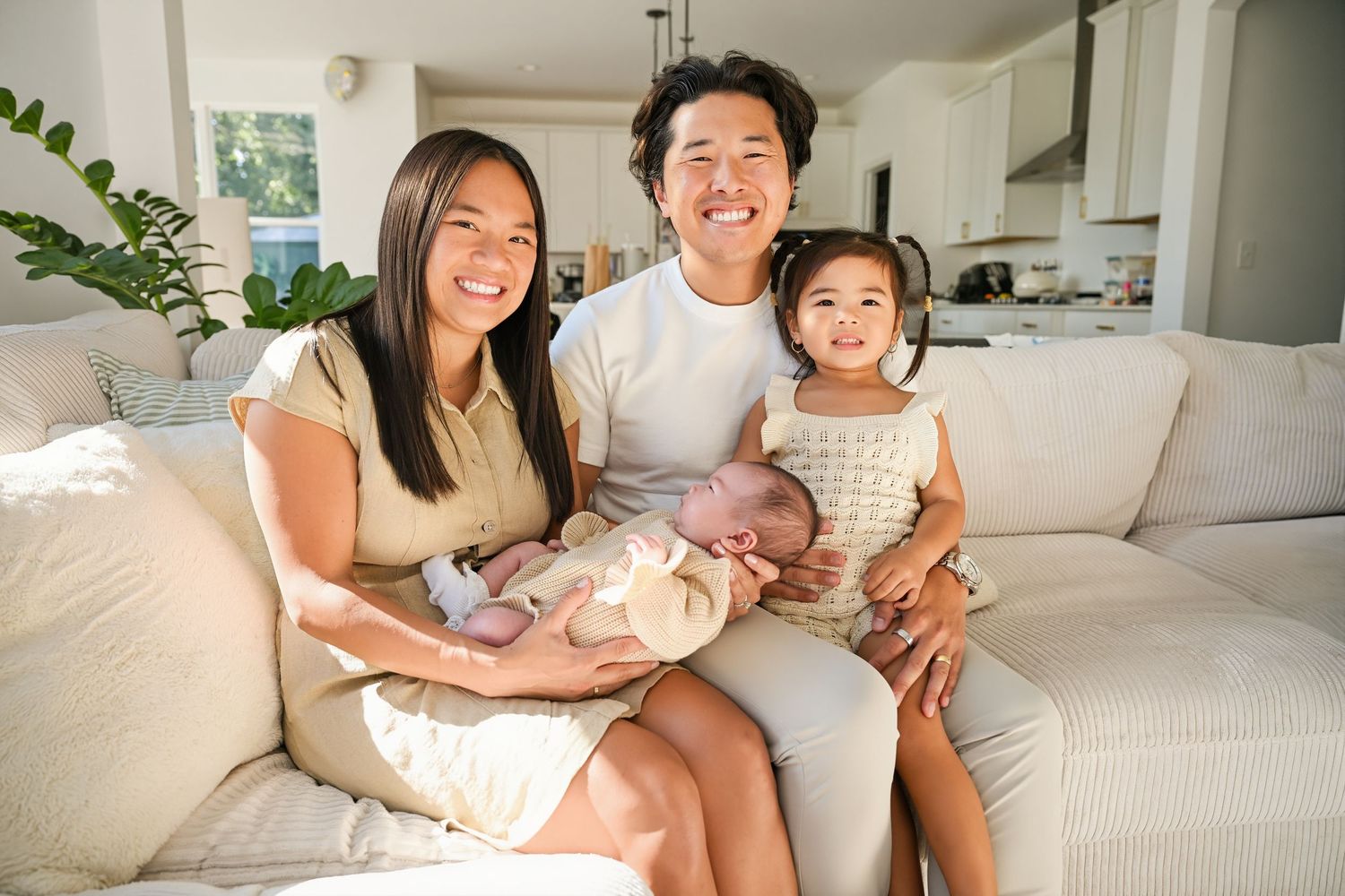 Joyful Asian family of four posing on white sofa, parents smiling with toddler girl and newborn baby in arms.
