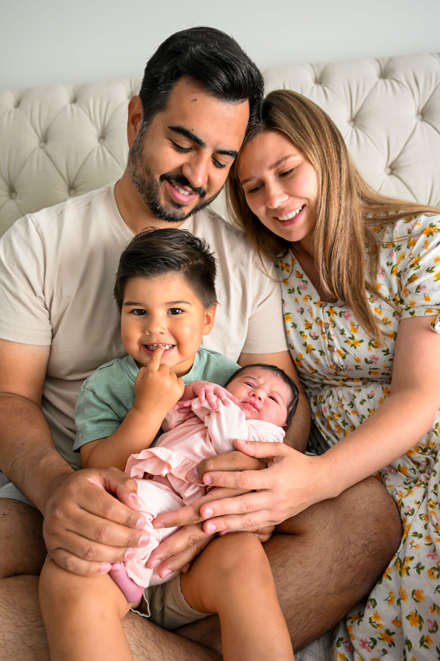 Smiling family of four with parents, toddler, and newborn baby sitting together on a bed.