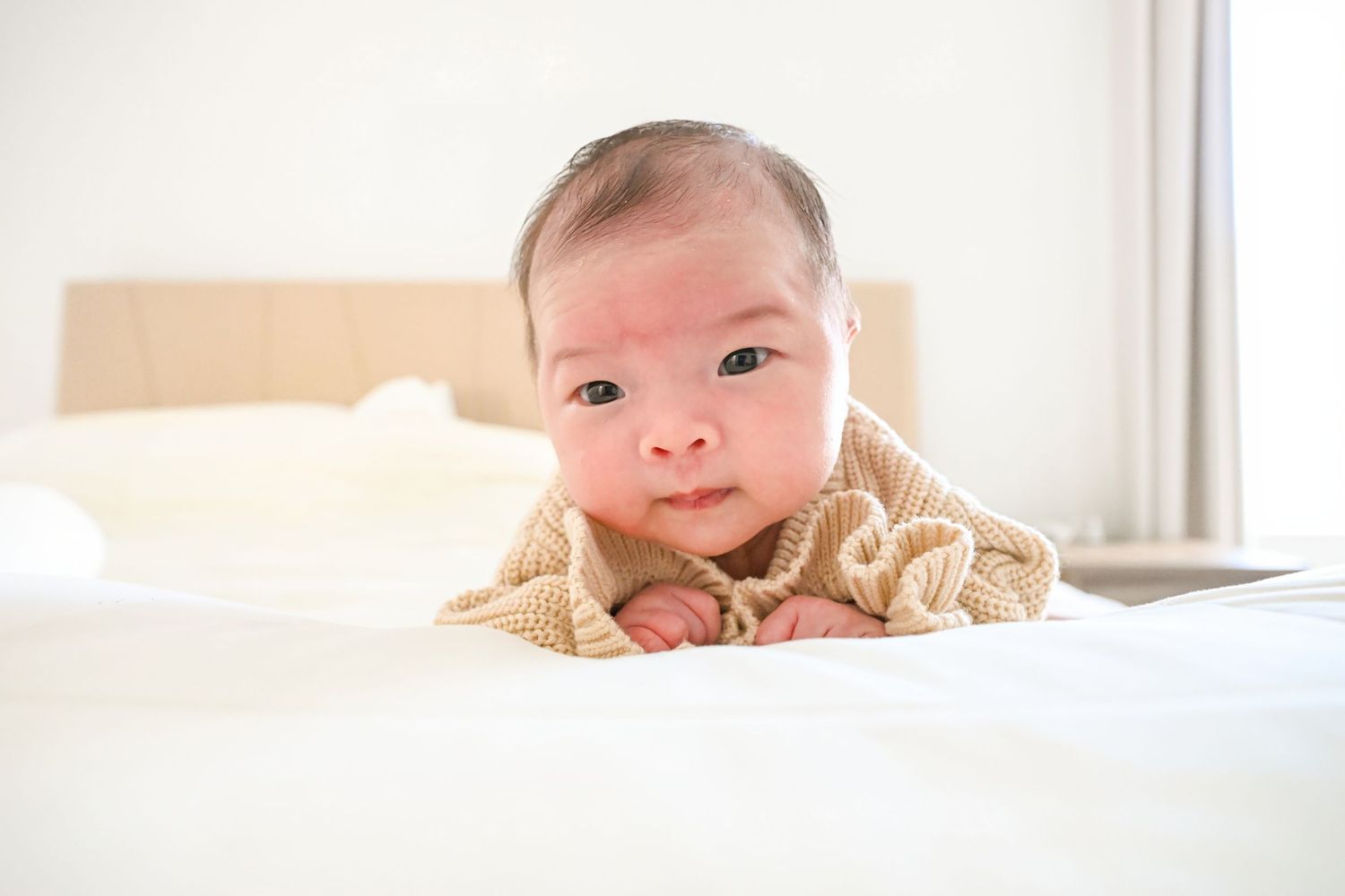 A cute Asian baby in a tan knit sweater lies on tummy on a white bed, looking curiously at the camera.