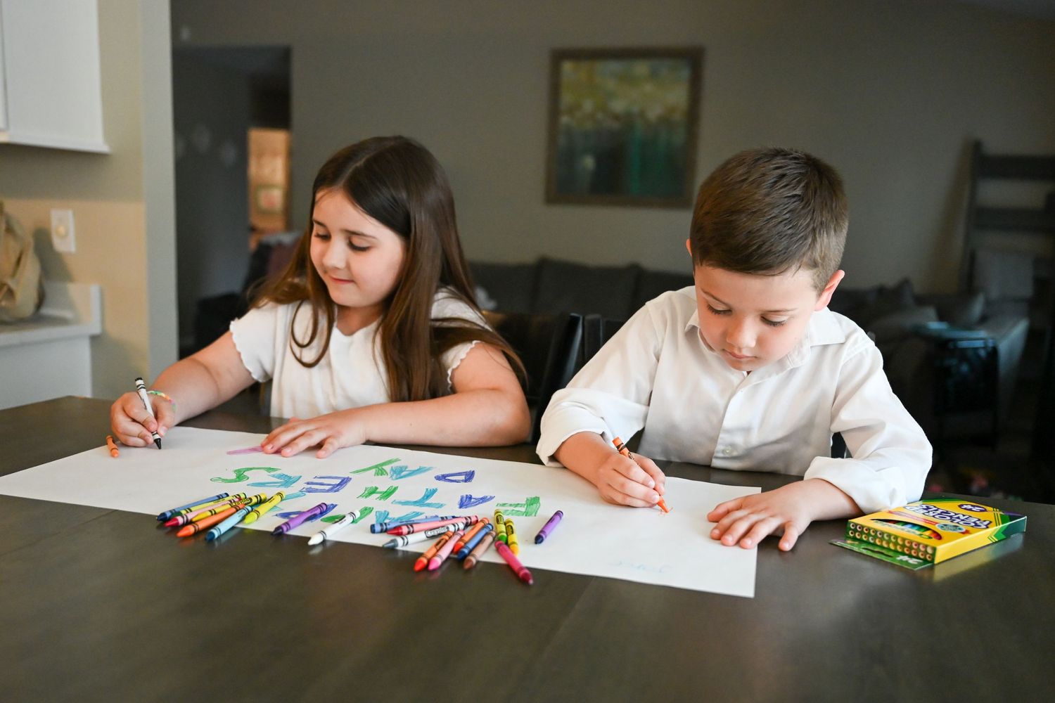 Two children sit at a dining table drawing and coloring on large white paper with Crayola crayons.