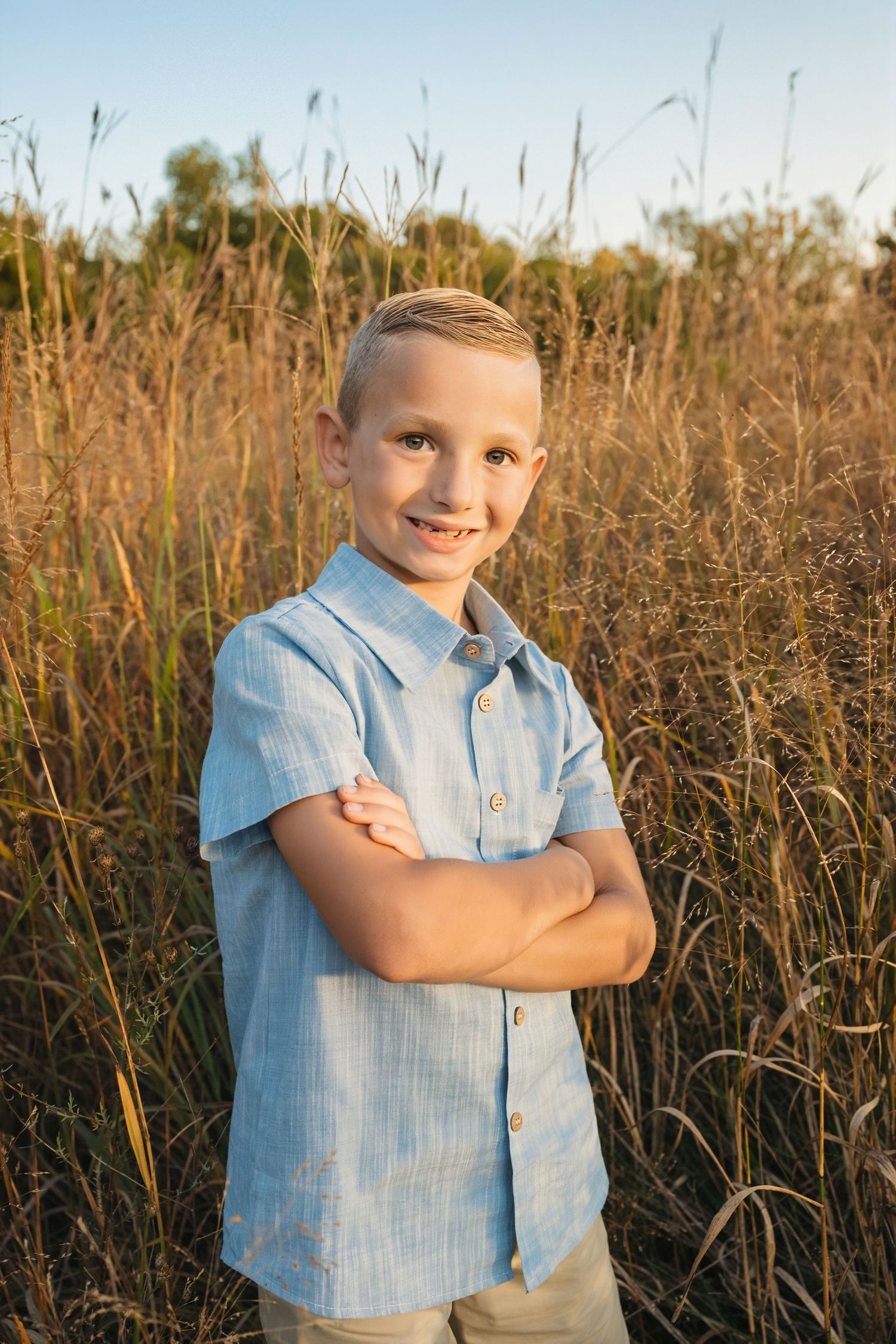 A smiling boy in a blue shirt stands with arms crossed in a golden field of tall grass during sunset.