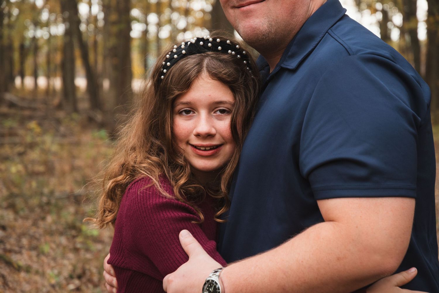 Young girl with headband hugging a man in a navy polo in a wooded fall setting.