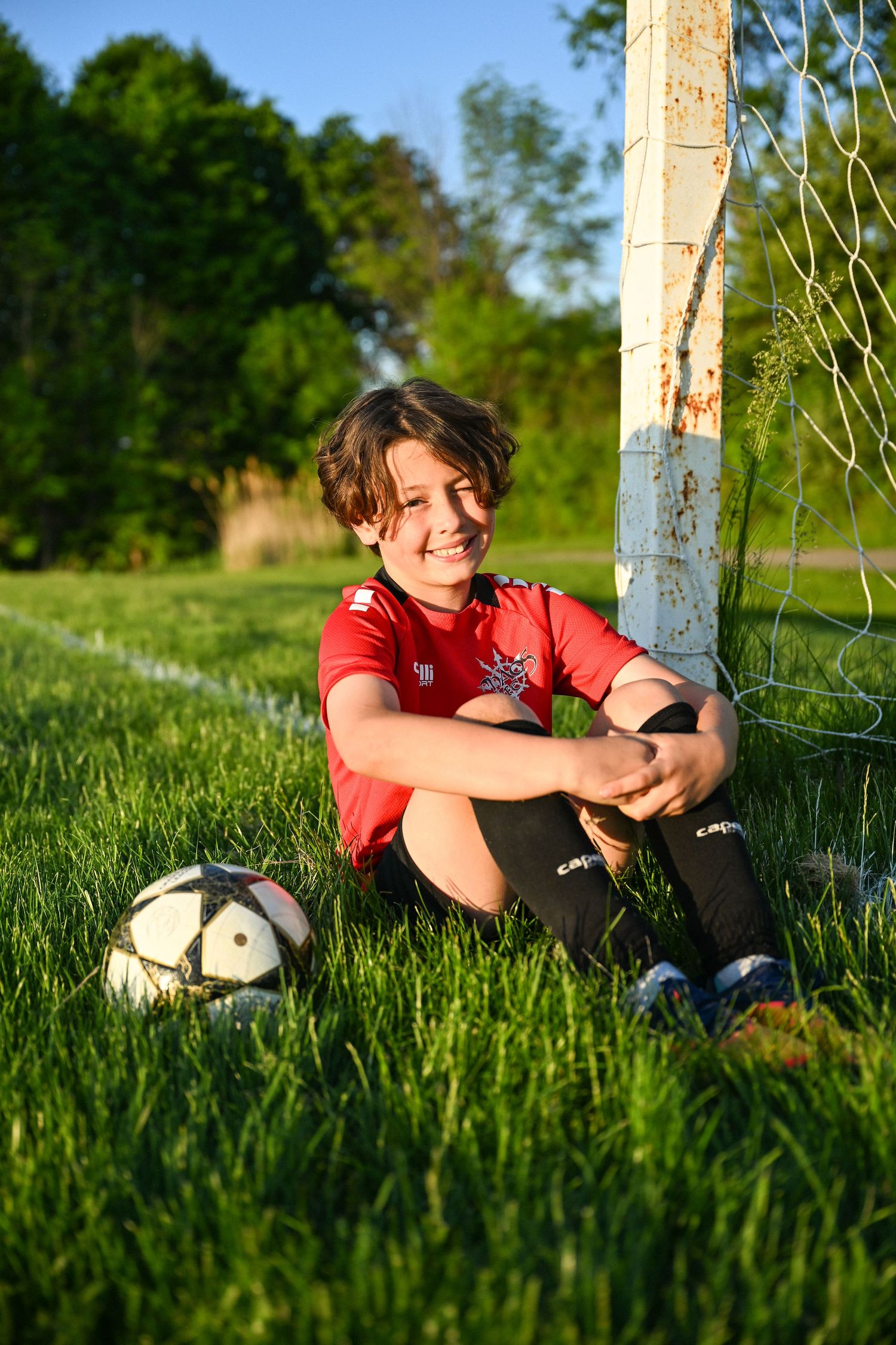 Smiling young soccer player in red uniform sitting on grass beside goal post with soccer ball on sunny day.