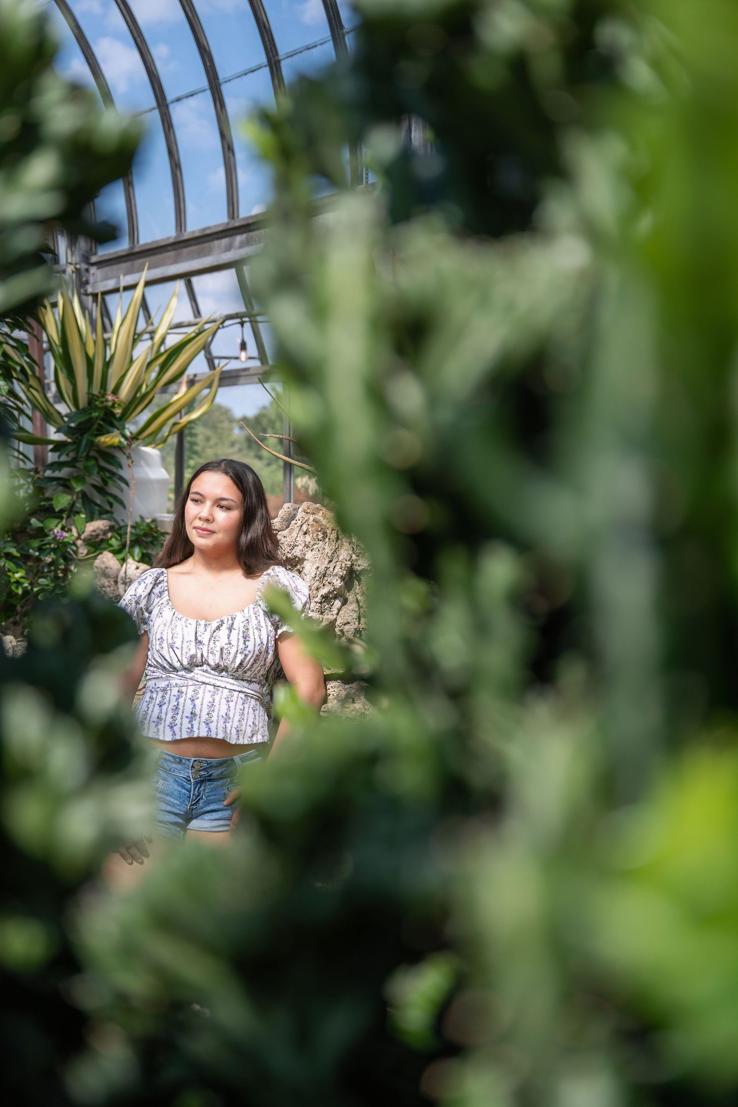 Teen girl in floral crop top and jeans photographed through lush green greenhouse plants.