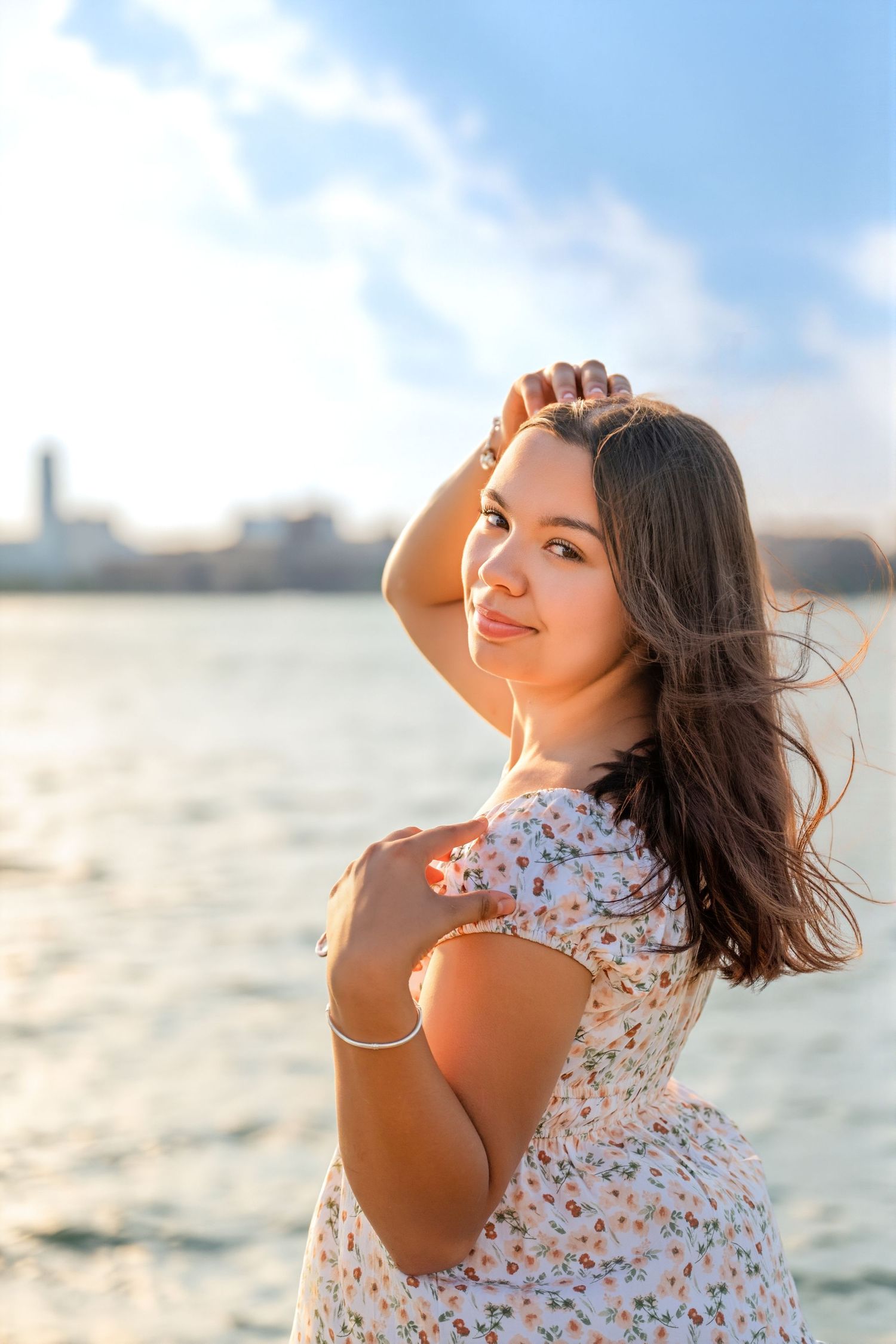 Young woman in floral dress posing by the waterfront at golden hour, hair blowing in the breeze.