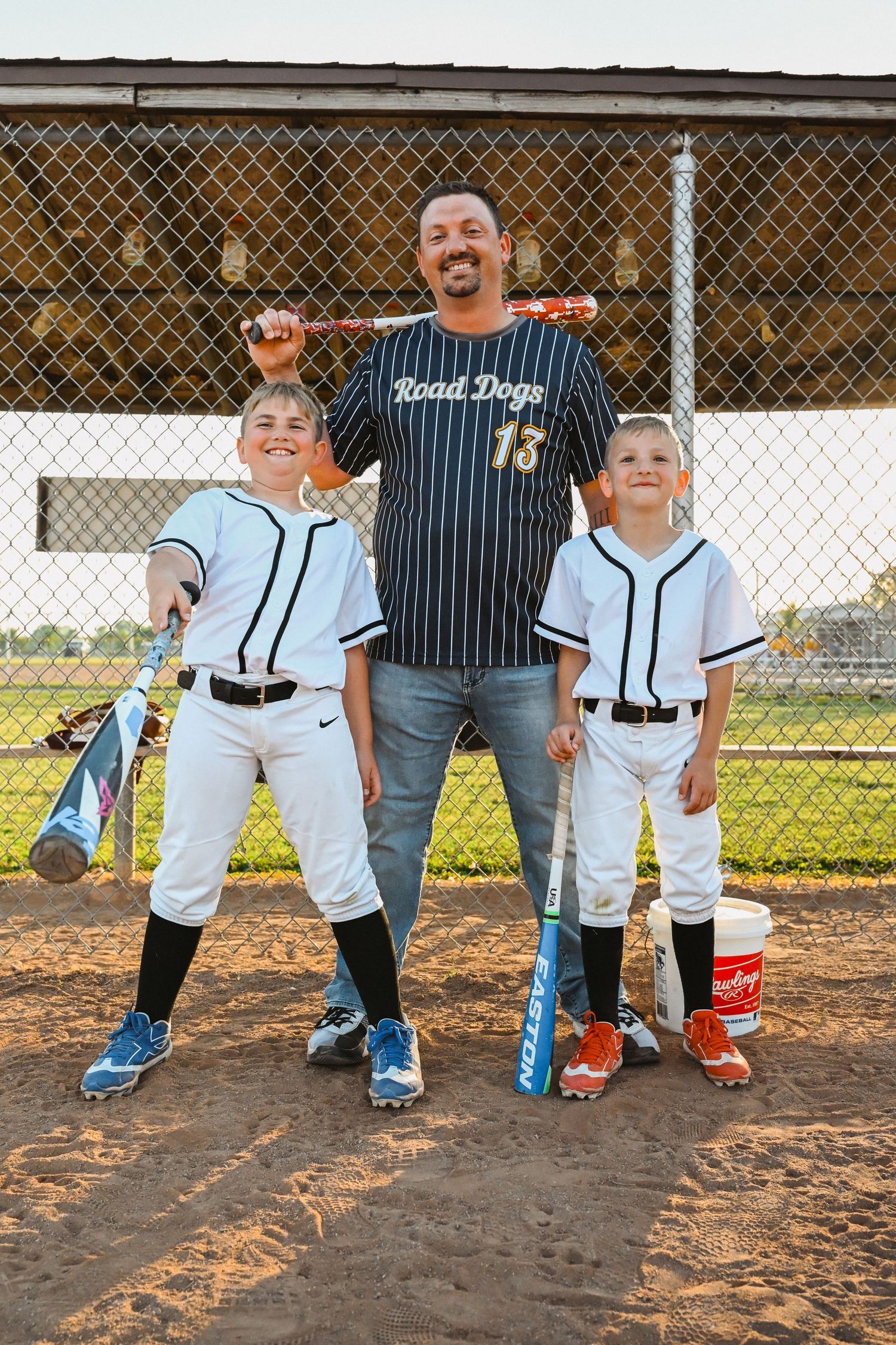 Father posing with two sons holding baseball bats near chain-link fence on field.