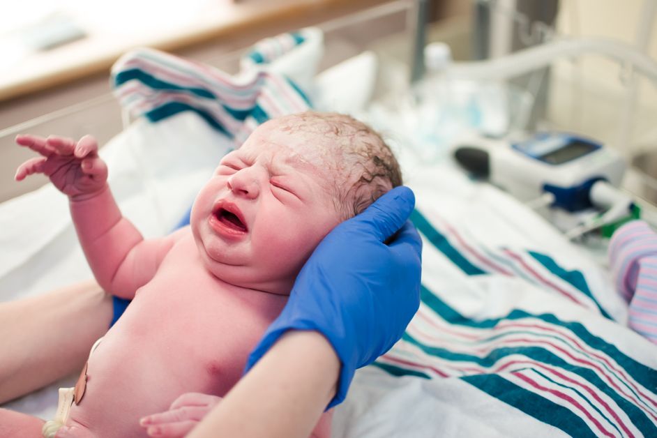 Crying newborn baby being examined by a medical professional wearing blue gloves at birth.