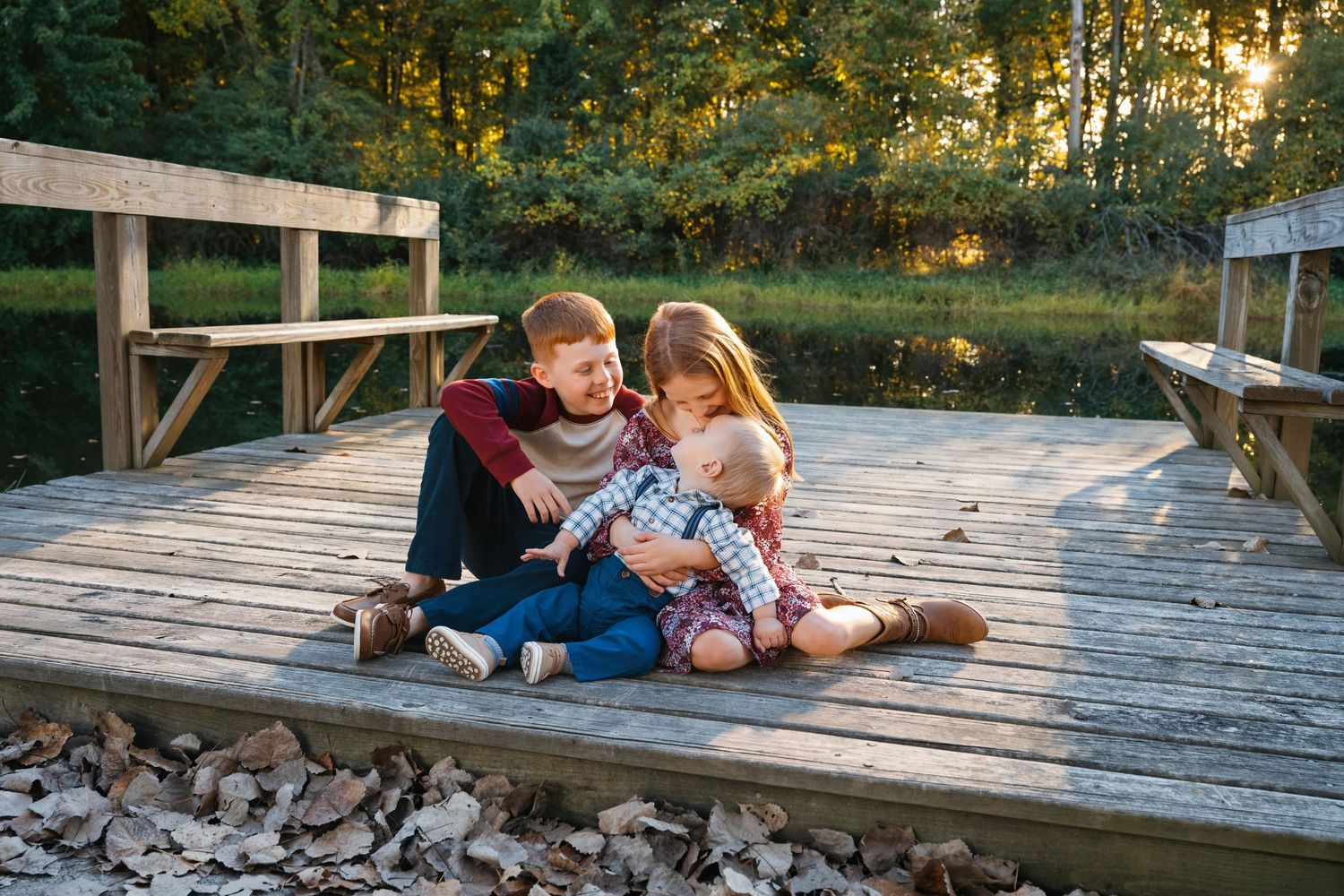 Three children sitting together on a wooden dock by a lake, enjoying a sunny autumn day outdoors.
