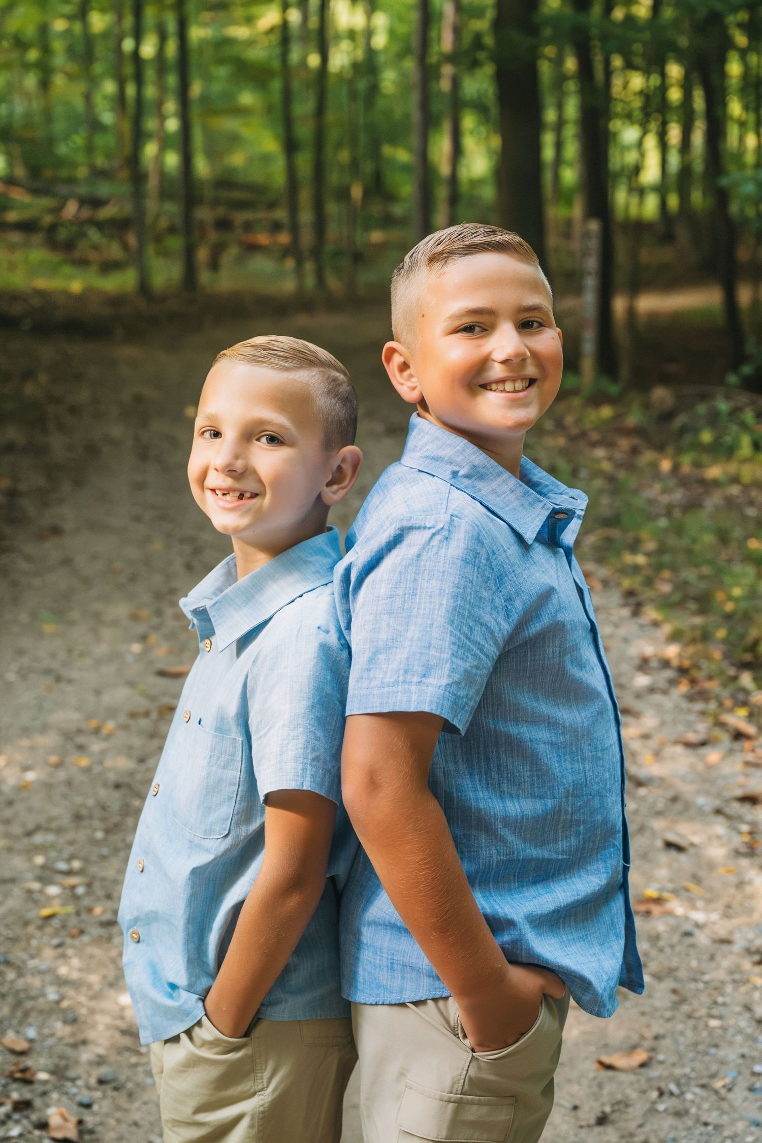 Two smiling boys in matching blue shirts stand back-to-back on a forest path, posing for a portrait outdoors.