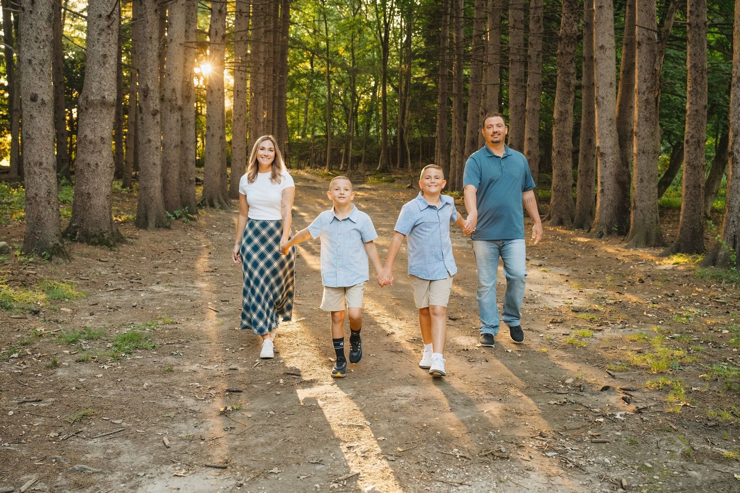 Family of four walking hand-in-hand on a sunlit forest path surrounded by tall pine trees.
