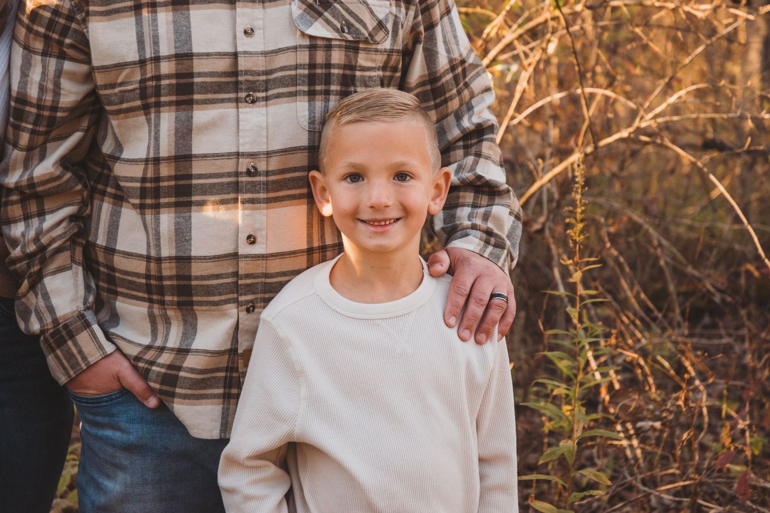 A smiling young boy in a cream sweatshirt stands outdoors in autumn with an adult in a plaid flannel shirt.