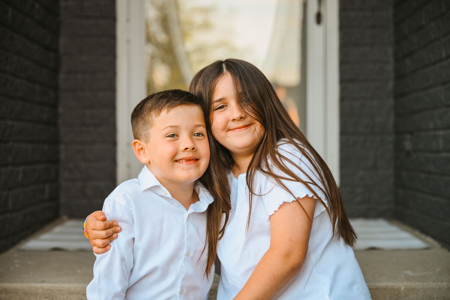 Two young siblings in white shirts embracing and smiling together on front porch steps.