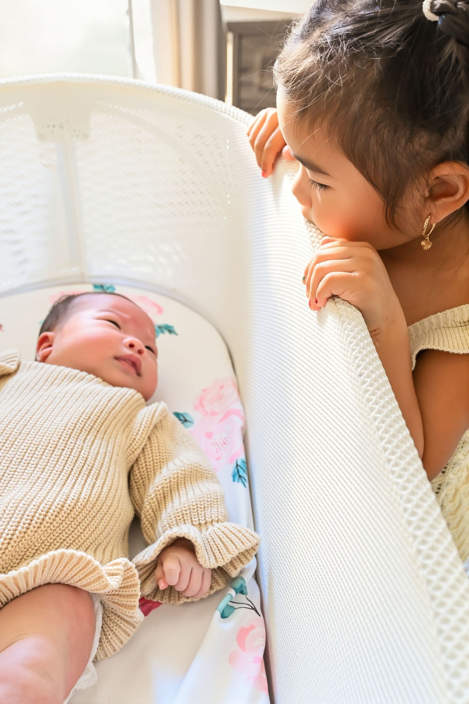 A young girl gazes lovingly at her newborn sibling lying in a white bassinet, wearing a cozy knit outfit.