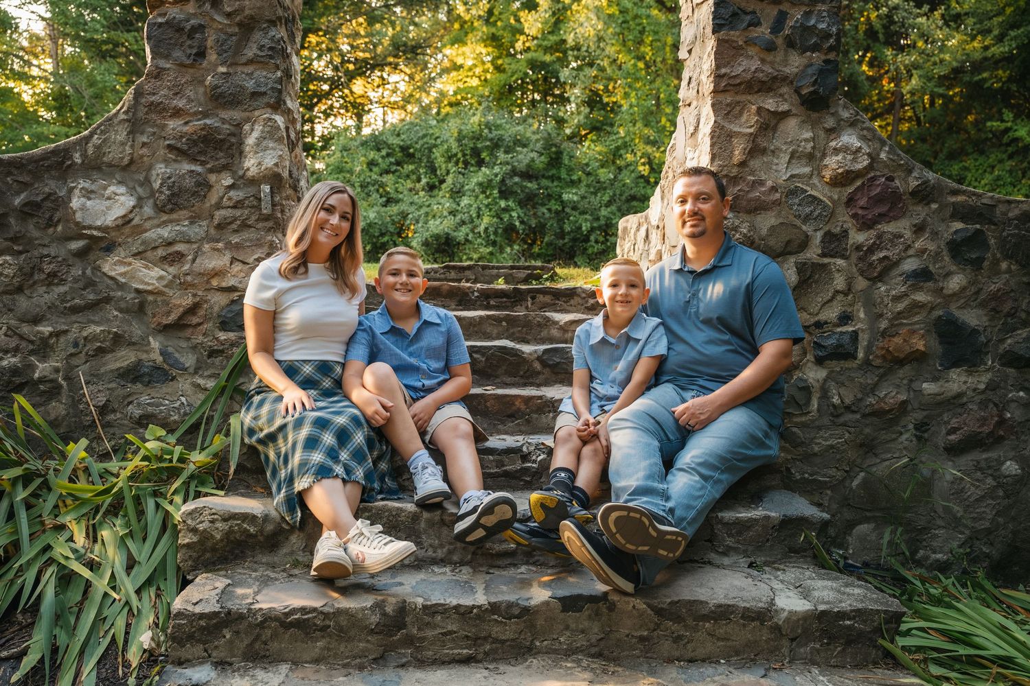 Family of four smiling on stone steps surrounded by lush greenery in an outdoor garden setting.