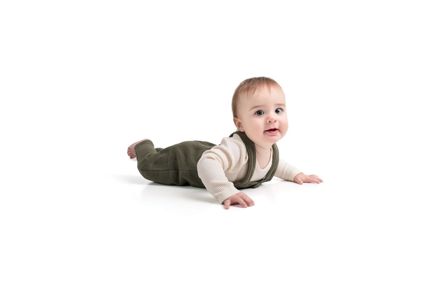 Cute baby in green overalls lying on stomach, smiling and looking up on white background.
