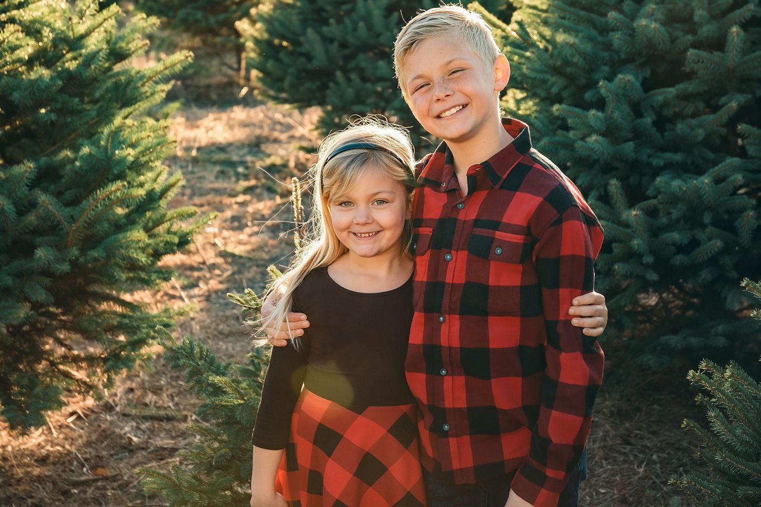 Two children in matching red plaid outfits smile together among Christmas trees at a holiday tree farm.
