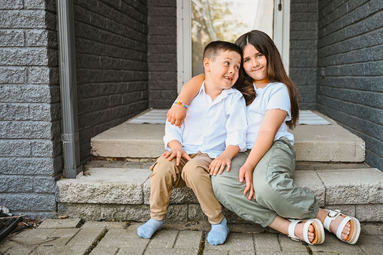 Two children sitting together on front porch steps smiling and hugging each other warmly.
