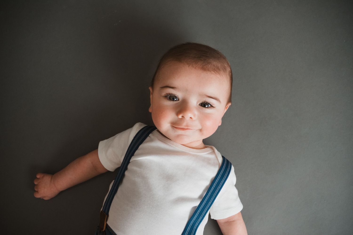 Adorable baby in white onesie with blue suspenders lying down and smiling at camera.